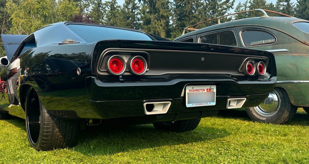 The rear view of a sleek black Dodge Charger, expertly restored by Horsepower NW. The car features modernized taillights and polished dual exhaust tips, highlighting its blend of vintage muscle car design and contemporary performance upgrades. The vehicle is parked on a grassy area at a car show, with a classic car visible in the background, showcasing the attention to detail and high-quality craftsmanship of Horsepower NW.