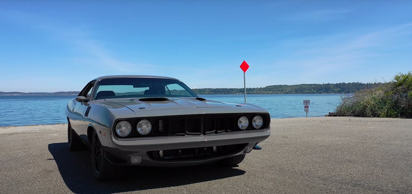 A meticulously restored grey Plymouth Barracuda parked on a coastal road, with a serene water backdrop and green hills. This ground-up custom build by Horsepower NW maintains its vintage soul while integrating modern performance and style enhancements, exemplifying a show-winning dream car built to be driven.
